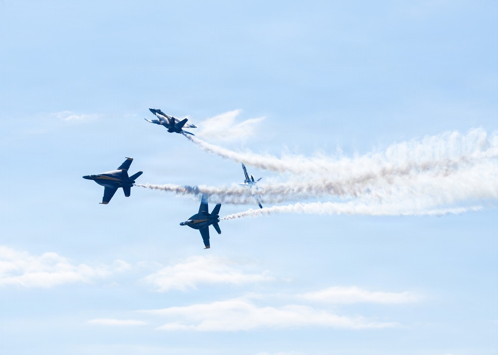 Four military jet aircraft are performing an aerobatic maneuver in a light blue sky, appearing to break apart or diverge from a central point. The jets are leaving white smoke trails that converge toward the right side of the frame, indicating a split or burst maneuver. The aircraft appear to be the U.S. Navy Blue Angels based on their dark blue coloring and F/A-18 Hornet silhouettes.