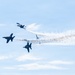 Four military jet aircraft are performing an aerobatic maneuver in a light blue sky, appearing to break apart or diverge from a central point. The jets are leaving white smoke trails that converge toward the right side of the frame, indicating a split or burst maneuver. The aircraft appear to be the U.S. Navy Blue Angels based on their dark blue coloring and F/A-18 Hornet silhouettes.