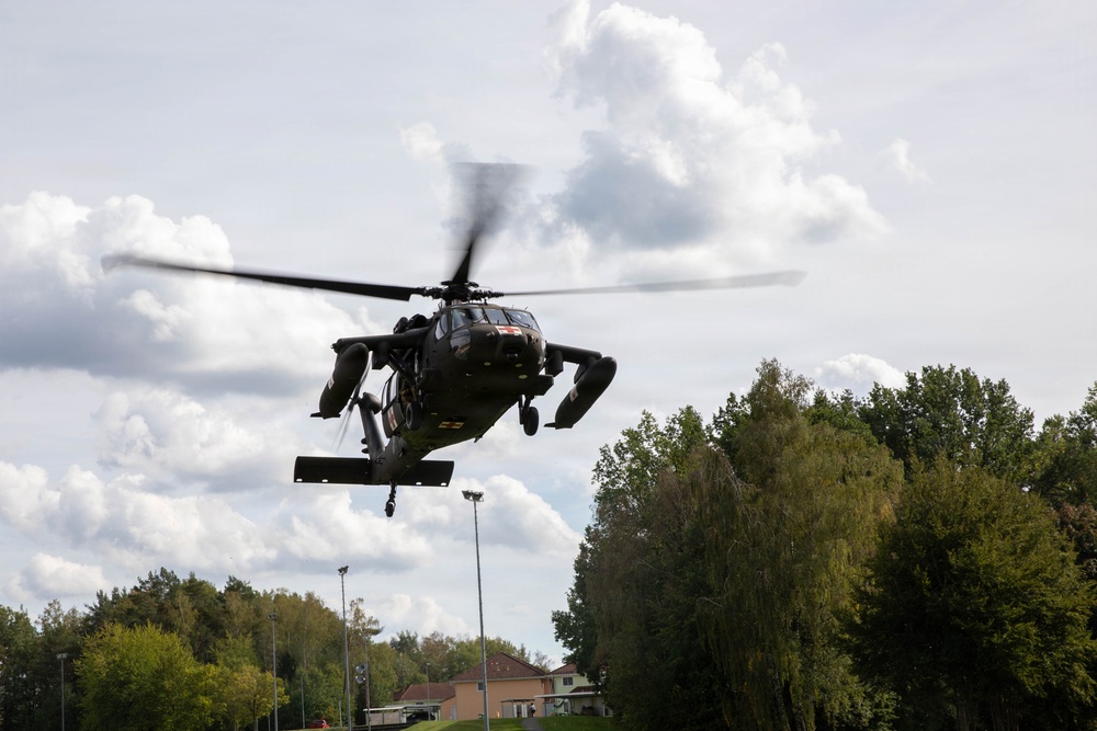 A military helicopter, consistent with a UH-60 Black Hawk design, is captured in low-altitude flight from a ground-level perspective. The helicopter appears to be in a hover or slow flight phase over what looks like a sports field or open area near a residential or base community. The rotor blades show motion blur indicating active flight.