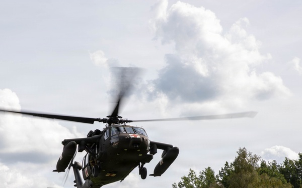A military helicopter, consistent with a UH-60 Black Hawk design, is captured in low-altitude flight from a ground-level perspective. The helicopter appears to be in a hover or slow flight phase over what looks like a sports field or open area near a residential or base community. The rotor blades show motion blur indicating active flight.