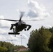 A military helicopter, consistent with a UH-60 Black Hawk design, is captured in low-altitude flight from a ground-level perspective. The helicopter appears to be in a hover or slow flight phase over what looks like a sports field or open area near a residential or base community. The rotor blades show motion blur indicating active flight.