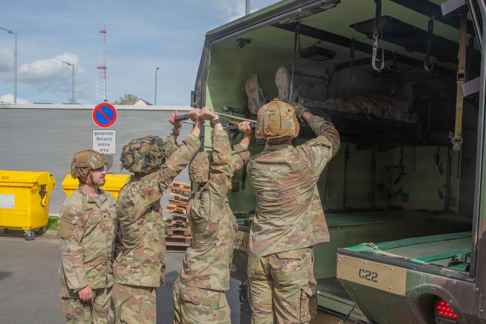 A group of U.S. Army soldiers in OCP (Operational Camouflage Pattern) uniforms and combat helmets are loading or unloading a litter/stretcher with a casualty into the rear of a military medical vehicle marked 'C22'. The scene appears to take place at a military installation in Germany, as evidenced by a German-language sign reading 'gesamter Bereich / entire area' alongside a no-parking road sign. This appears to be a medical evacuation (MEDEVAC) training exercise or actual casualty transport operation.