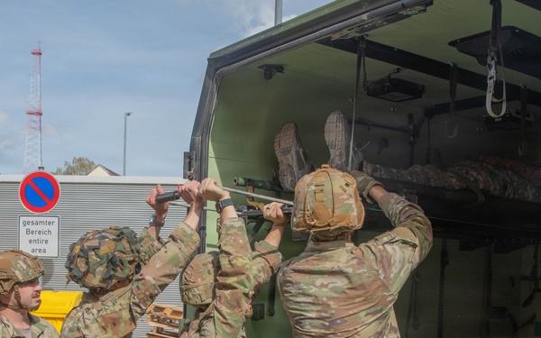 A group of U.S. Army soldiers in OCP (Operational Camouflage Pattern) uniforms and combat helmets are loading or unloading a litter/stretcher with a casualty into the rear of a military medical vehicle marked 'C22'. The scene appears to take place at a military installation in Germany, as evidenced by a German-language sign reading 'gesamter Bereich / entire area' alongside a no-parking road sign. This appears to be a medical evacuation (MEDEVAC) training exercise or actual casualty transport operation.