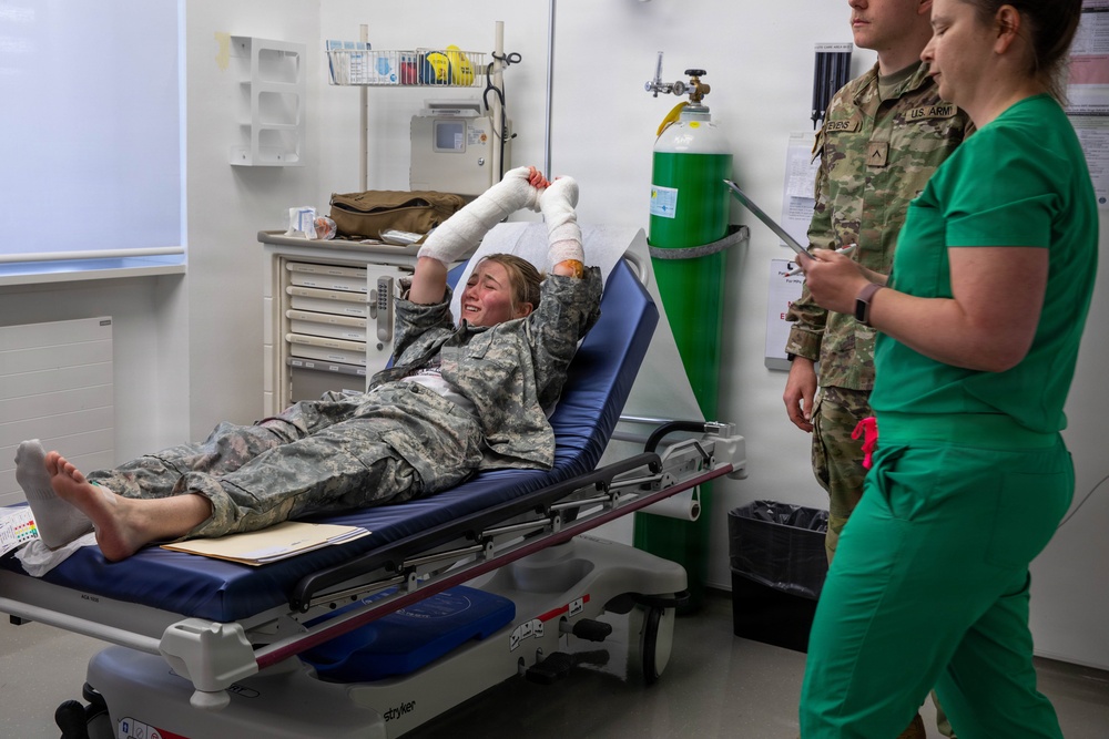 A military personnel member in Army Combat Uniform (ACU) is lying on a Stryker medical gurney with both hands heavily bandaged, appearing to be in pain or distress. Two medical/military staff are present — one in U.S. Army uniform and one in green medical scrubs holding a tablet or clipboard. The setting appears to be a military medical treatment facility or emergency room.