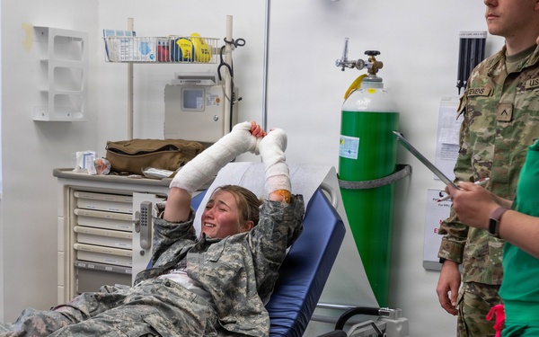 A military personnel member in Army Combat Uniform (ACU) is lying on a Stryker medical gurney with both hands heavily bandaged, appearing to be in pain or distress. Two medical/military staff are present — one in U.S. Army uniform and one in green medical scrubs holding a tablet or clipboard. The setting appears to be a military medical treatment facility or emergency room.