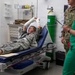 A military personnel member in Army Combat Uniform (ACU) is lying on a Stryker medical gurney with both hands heavily bandaged, appearing to be in pain or distress. Two medical/military staff are present — one in U.S. Army uniform and one in green medical scrubs holding a tablet or clipboard. The setting appears to be a military medical treatment facility or emergency room.