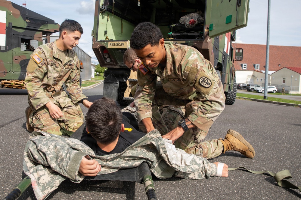 Three U.S. Army soldiers in OCP (Operational Camouflage Pattern) uniforms are conducting a casualty evacuation drill near a military vehicle marked 'RSS/2CAV'. One soldier is acting as a simulated casualty lying on a litter/stretcher on the pavement, while two others work to load or position him. The scene appears to be a training exercise at a military installation, likely in Europe based on the architecture.