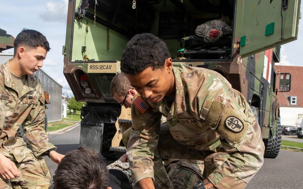 Three U.S. Army soldiers in OCP (Operational Camouflage Pattern) uniforms are conducting a casualty evacuation drill near a military vehicle marked 'RSS/2CAV'. One soldier is acting as a simulated casualty lying on a litter/stretcher on the pavement, while two others work to load or position him. The scene appears to be a training exercise at a military installation, likely in Europe based on the architecture.