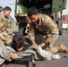 Three U.S. Army soldiers in OCP (Operational Camouflage Pattern) uniforms are conducting a casualty evacuation drill near a military vehicle marked 'RSS/2CAV'. One soldier is acting as a simulated casualty lying on a litter/stretcher on the pavement, while two others work to load or position him. The scene appears to be a training exercise at a military installation, likely in Europe based on the architecture.