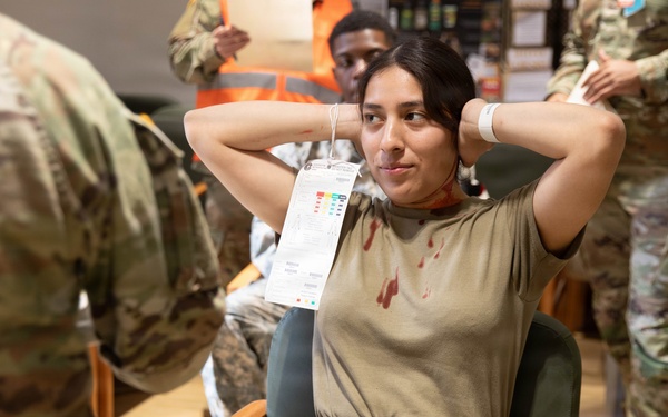 A military training exercise or medical drill is depicted, showing a female soldier seated in a chair with simulated blood stains on her uniform and a medical triage tag hanging from her neck. She has her hands clasped behind her head in a relaxed posture, suggesting she is acting as a simulated casualty. Several other personnel in military uniforms and at least one person in an orange safety vest are visible in the background.