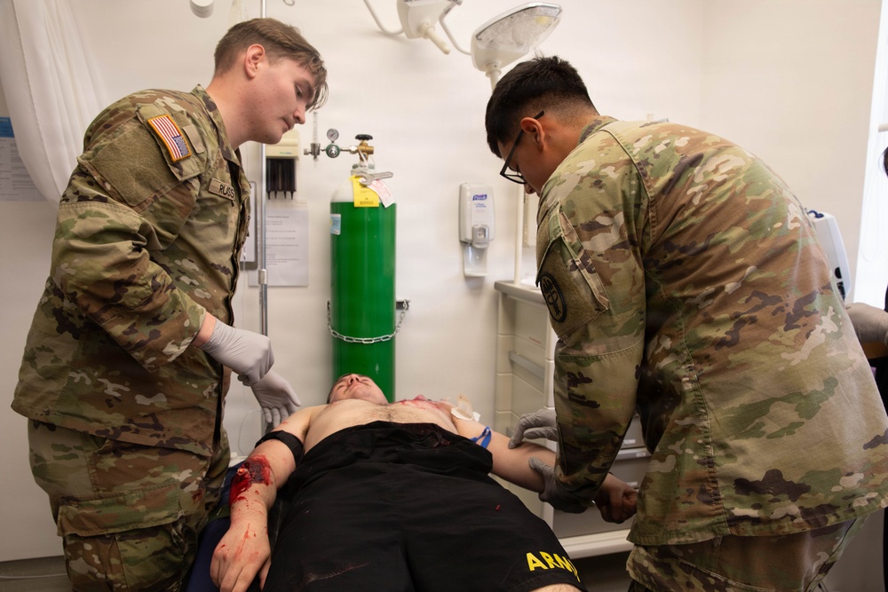 Two U.S. Army soldiers in OCP (Operational Camouflage Pattern) uniforms are providing medical treatment to a casualty lying on an examination table. The scene appears to be a medical training exercise or simulation, indicated by the controlled clinical environment and simulated wounds visible on the patient. Both soldiers are wearing gloves and actively working on the patient's arms and torso.