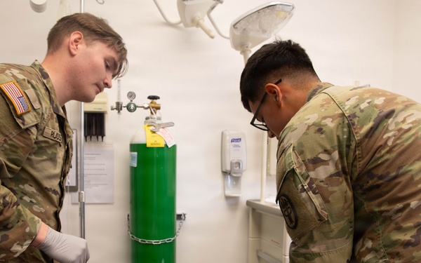 Two U.S. Army soldiers in OCP (Operational Camouflage Pattern) uniforms are providing medical treatment to a casualty lying on an examination table. The scene appears to be a medical training exercise or simulation, indicated by the controlled clinical environment and simulated wounds visible on the patient. Both soldiers are wearing gloves and actively working on the patient's arms and torso.