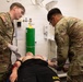 Two U.S. Army soldiers in OCP (Operational Camouflage Pattern) uniforms are providing medical treatment to a casualty lying on an examination table. The scene appears to be a medical training exercise or simulation, indicated by the controlled clinical environment and simulated wounds visible on the patient. Both soldiers are wearing gloves and actively working on the patient's arms and torso.