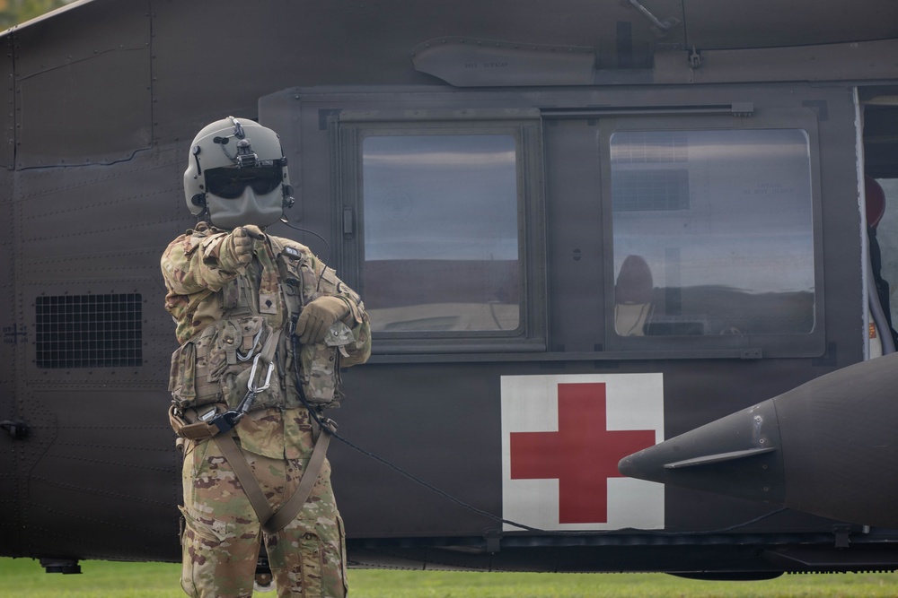 A military personnel in full combat gear and flight helmet stands facing a military medical evacuation (MEDEVAC) helicopter, identifiable by the prominent Red Cross symbol on its fuselage. The individual appears to be communicating with or signaling the aircraft crew through the cockpit window. The scene takes place on a grassy area, suggesting a landing zone or field operation.