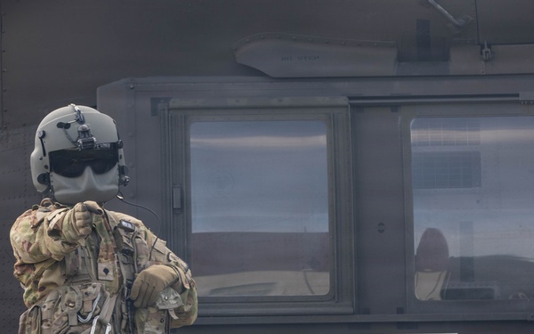 A military personnel in full combat gear and flight helmet stands facing a military medical evacuation (MEDEVAC) helicopter, identifiable by the prominent Red Cross symbol on its fuselage. The individual appears to be communicating with or signaling the aircraft crew through the cockpit window. The scene takes place on a grassy area, suggesting a landing zone or field operation.