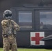 A military personnel in full combat gear and flight helmet stands facing a military medical evacuation (MEDEVAC) helicopter, identifiable by the prominent Red Cross symbol on its fuselage. The individual appears to be communicating with or signaling the aircraft crew through the cockpit window. The scene takes place on a grassy area, suggesting a landing zone or field operation.