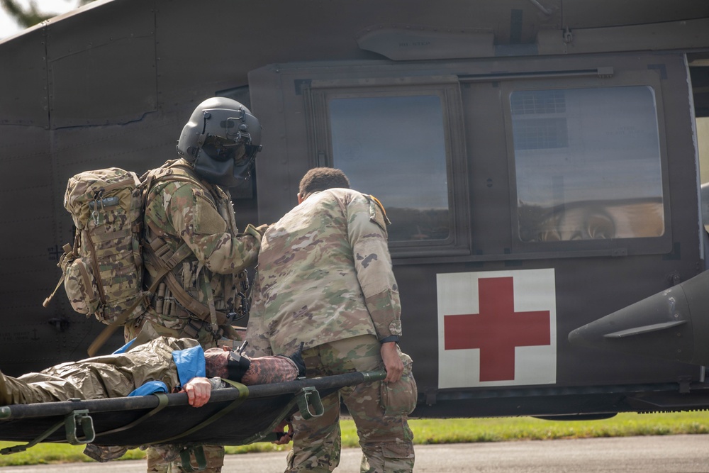 Two military personnel in camouflage uniforms are carrying a casualty on a stretcher toward a military medical evacuation (MEDEVAC) helicopter marked with a red cross symbol. One soldier is wearing a flight helmet and carrying a large tactical backpack, while the other assists in transporting the litter patient. This appears to be either a real MEDEVAC operation or a military medical training exercise.