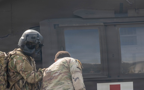 Two military personnel in camouflage uniforms are carrying a casualty on a stretcher toward a military medical evacuation (MEDEVAC) helicopter marked with a red cross symbol. One soldier is wearing a flight helmet and carrying a large tactical backpack, while the other assists in transporting the litter patient. This appears to be either a real MEDEVAC operation or a military medical training exercise.
