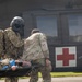 Two military personnel in camouflage uniforms are carrying a casualty on a stretcher toward a military medical evacuation (MEDEVAC) helicopter marked with a red cross symbol. One soldier is wearing a flight helmet and carrying a large tactical backpack, while the other assists in transporting the litter patient. This appears to be either a real MEDEVAC operation or a military medical training exercise.
