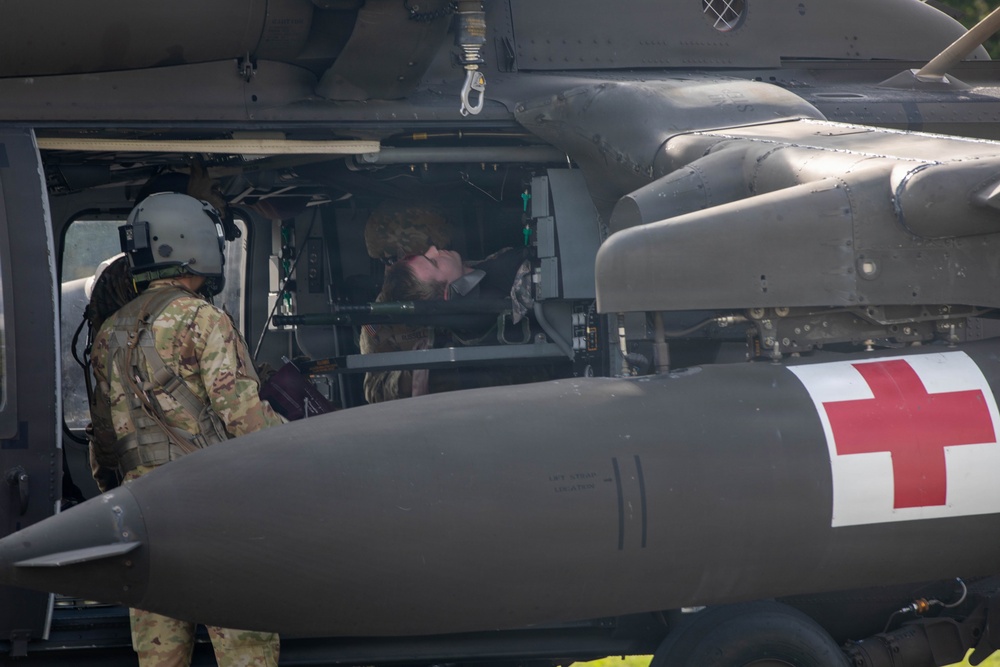 The image shows a military medical evacuation (MEDEVAC) helicopter, identifiable by the prominent red cross symbol on its external fuel tank or sponson. Two personnel in military camouflage uniforms are visible, one wearing a flight helmet standing outside the aircraft and another inside the cabin. The helicopter appears to be an olive-drab colored rotary-wing aircraft consistent with a UH-60 Black Hawk variant configured for medical operations.