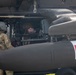 The image shows a military medical evacuation (MEDEVAC) helicopter, identifiable by the prominent red cross symbol on its external fuel tank or sponson. Two personnel in military camouflage uniforms are visible, one wearing a flight helmet standing outside the aircraft and another inside the cabin. The helicopter appears to be an olive-drab colored rotary-wing aircraft consistent with a UH-60 Black Hawk variant configured for medical operations.