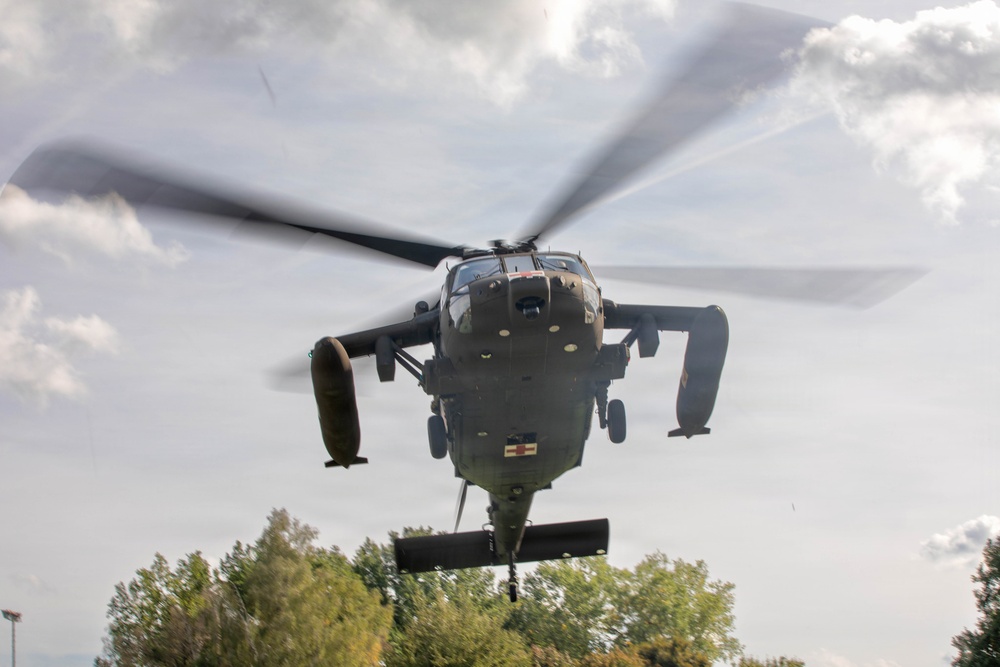 A military helicopter is captured in a low-altitude hover or landing approach, photographed from a front-facing perspective. The helicopter displays a red cross marking on its fuselage, indicating a medical/medevac role. The rotor blades are visibly motion-blurred, suggesting active flight, with trees and a partly cloudy sky visible in the background.