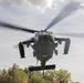 A military helicopter is captured in a low-altitude hover or landing approach, photographed from a front-facing perspective. The helicopter displays a red cross marking on its fuselage, indicating a medical/medevac role. The rotor blades are visibly motion-blurred, suggesting active flight, with trees and a partly cloudy sky visible in the background.