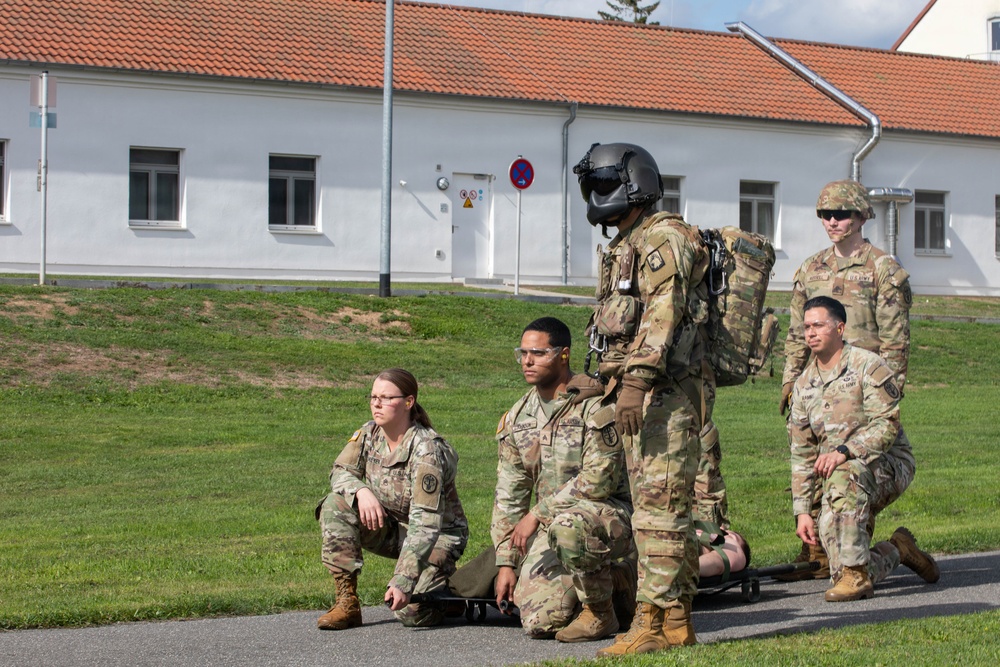 A group of five U.S. Army soldiers in MultiCam (OCP) uniforms are gathered on a paved area adjacent to a grass field, in front of a white building with a red-tiled roof. One soldier is wearing a flight helmet and appears to be equipped with a flight suit and harness, suggesting aviation-related training or a demonstration. The soldiers appear to be in a waiting or observing posture, possibly during a training exercise.