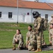 A group of five U.S. Army soldiers in MultiCam (OCP) uniforms are gathered on a paved area adjacent to a grass field, in front of a white building with a red-tiled roof. One soldier is wearing a flight helmet and appears to be equipped with a flight suit and harness, suggesting aviation-related training or a demonstration. The soldiers appear to be in a waiting or observing posture, possibly during a training exercise.
