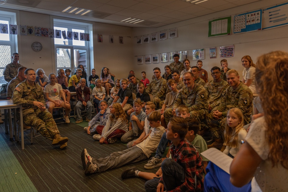 A group of U.S. Army soldiers in camouflage uniforms are visiting a school classroom, seated alongside a large group of children. A woman with curly hair, likely a teacher, is addressing the group while holding papers. The setting appears to be a European school based on the classroom decor and architectural style.