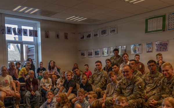 A group of U.S. Army soldiers in camouflage uniforms are visiting a school classroom, seated alongside a large group of children. A woman with curly hair, likely a teacher, is addressing the group while holding papers. The setting appears to be a European school based on the classroom decor and architectural style.