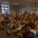 A group of U.S. Army soldiers in camouflage uniforms are visiting a school classroom, seated alongside a large group of children. A woman with curly hair, likely a teacher, is addressing the group while holding papers. The setting appears to be a European school based on the classroom decor and architectural style.