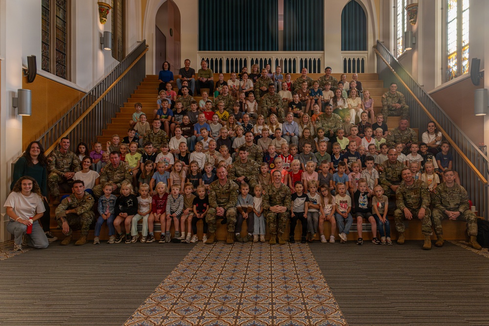 A large group photograph taken inside what appears to be a historic chapel or church building, featuring numerous military personnel in camouflage uniforms posed on a grand staircase alongside many children and a few civilians. The group spans multiple rows across wide wooden stairs, suggesting a formal unit or family day event. Stained glass windows and ornate architectural details are visible in the background.