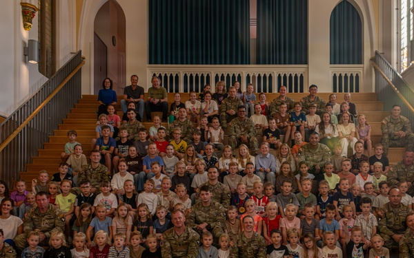 A large group photograph taken inside what appears to be a historic chapel or church building, featuring numerous military personnel in camouflage uniforms posed on a grand staircase alongside many children and a few civilians. The group spans multiple rows across wide wooden stairs, suggesting a formal unit or family day event. Stained glass windows and ornate architectural details are visible in the background.