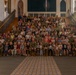 A large group photograph taken inside what appears to be a historic chapel or church building, featuring numerous military personnel in camouflage uniforms posed on a grand staircase alongside many children and a few civilians. The group spans multiple rows across wide wooden stairs, suggesting a formal unit or family day event. Stained glass windows and ornate architectural details are visible in the background.