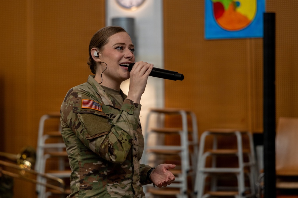 A female U.S. Army soldier in Operational Camouflage Pattern (OCP) uniform is singing into a handheld microphone while wearing an in-ear monitor. She appears to be performing at an indoor venue, likely a school or community center, as suggested by the colorful artwork visible in the background. A brass instrument, possibly a trombone, is partially visible in the lower left corner.
