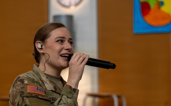 A female U.S. Army soldier in Operational Camouflage Pattern (OCP) uniform is singing into a handheld microphone while wearing an in-ear monitor. She appears to be performing at an indoor venue, likely a school or community center, as suggested by the colorful artwork visible in the background. A brass instrument, possibly a trombone, is partially visible in the lower left corner.