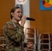 A female U.S. Army soldier in Operational Camouflage Pattern (OCP) uniform is singing into a handheld microphone while wearing an in-ear monitor. She appears to be performing at an indoor venue, likely a school or community center, as suggested by the colorful artwork visible in the background. A brass instrument, possibly a trombone, is partially visible in the lower left corner.