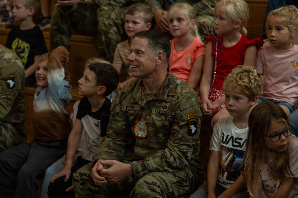 A U.S. Army soldier named REIM, wearing OCP (Operational Camouflage Pattern) uniform with 101st Airborne Division patch and Ranger/Airborne tabs, sits among a group of young children in what appears to be a school or community gathering. The soldier is smiling and appears engaged with the children around him. The setting suggests a military community outreach or school visit event.