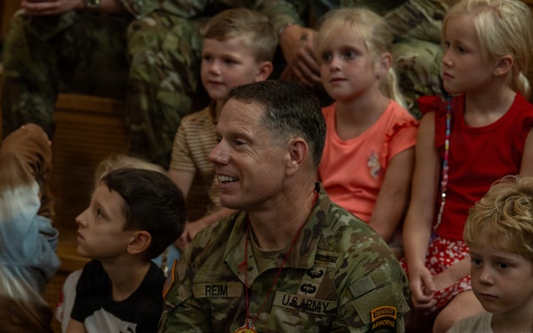 A U.S. Army soldier named REIM, wearing OCP (Operational Camouflage Pattern) uniform with 101st Airborne Division patch and Ranger/Airborne tabs, sits among a group of young children in what appears to be a school or community gathering. The soldier is smiling and appears engaged with the children around him. The setting suggests a military community outreach or school visit event.