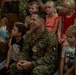 A U.S. Army soldier named REIM, wearing OCP (Operational Camouflage Pattern) uniform with 101st Airborne Division patch and Ranger/Airborne tabs, sits among a group of young children in what appears to be a school or community gathering. The soldier is smiling and appears engaged with the children around him. The setting suggests a military community outreach or school visit event.