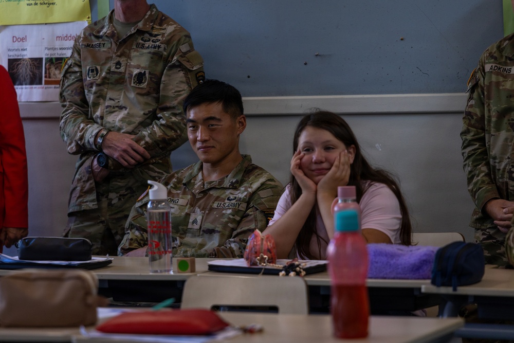 U.S. Army soldiers are visiting a classroom, likely in a Dutch-speaking country based on the visible Dutch text on educational posters. A seated soldier with the name tag 'ONG' (partially visible) sits alongside a young female student at a school desk. At least two other soldiers, identified by name tags 'MASSEY' and 'ADKINS', are also present in the room.