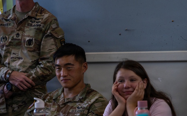 U.S. Army soldiers are visiting a classroom, likely in a Dutch-speaking country based on the visible Dutch text on educational posters. A seated soldier with the name tag 'ONG' (partially visible) sits alongside a young female student at a school desk. At least two other soldiers, identified by name tags 'MASSEY' and 'ADKINS', are also present in the room.
