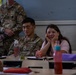 U.S. Army soldiers are visiting a classroom, likely in a Dutch-speaking country based on the visible Dutch text on educational posters. A seated soldier with the name tag 'ONG' (partially visible) sits alongside a young female student at a school desk. At least two other soldiers, identified by name tags 'MASSEY' and 'ADKINS', are also present in the room.