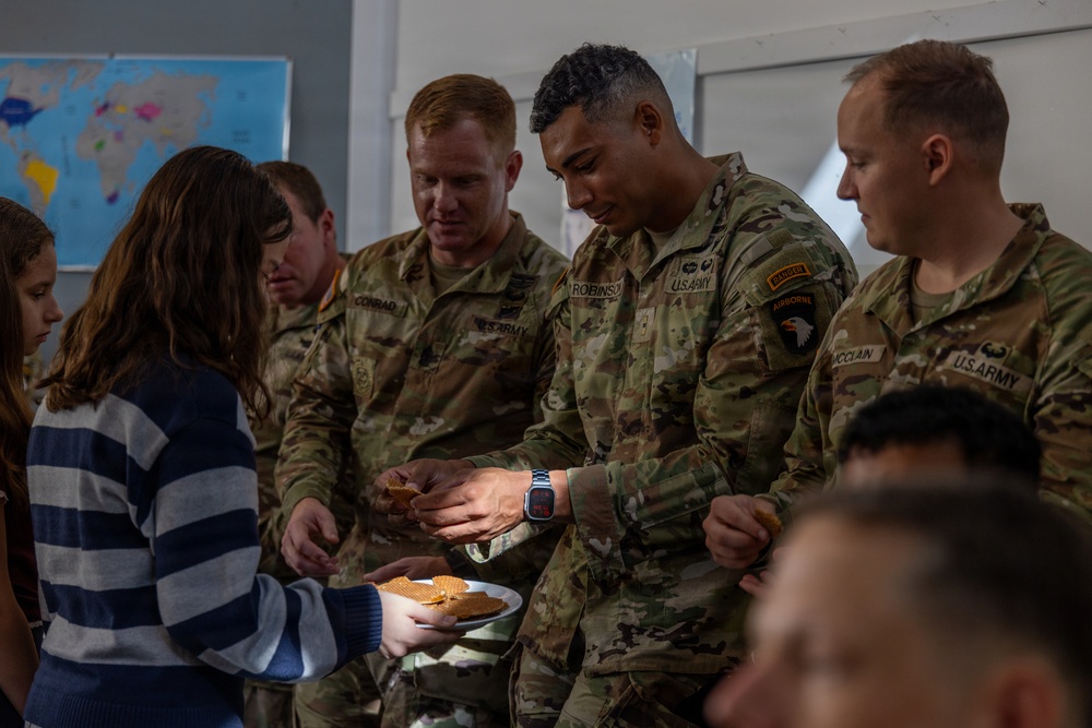 Several U.S. Army soldiers in OCP (Operational Camouflage Pattern) uniforms interact with a civilian, appearing to share or receive food items that look like waffles or waffle cookies. The setting appears to be an indoor community or educational space, evidenced by a world map poster visible on the wall in the background. The interaction appears friendly and informal, suggesting a community engagement or goodwill visit.