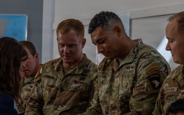 Several U.S. Army soldiers in OCP (Operational Camouflage Pattern) uniforms interact with a civilian, appearing to share or receive food items that look like waffles or waffle cookies. The setting appears to be an indoor community or educational space, evidenced by a world map poster visible on the wall in the background. The interaction appears friendly and informal, suggesting a community engagement or goodwill visit.