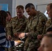 Several U.S. Army soldiers in OCP (Operational Camouflage Pattern) uniforms interact with a civilian, appearing to share or receive food items that look like waffles or waffle cookies. The setting appears to be an indoor community or educational space, evidenced by a world map poster visible on the wall in the background. The interaction appears friendly and informal, suggesting a community engagement or goodwill visit.