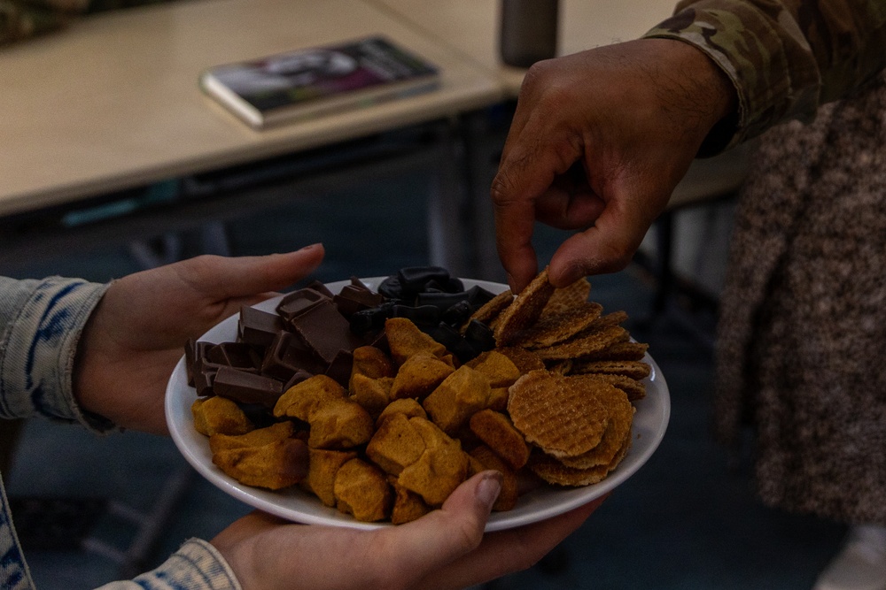 Two people are sharing a plate of assorted snacks, with one person holding the plate and another reaching to take a chip. The plate contains a variety of snacks including chocolate pieces, crackers or waffle chips, and what appear to be cheese puffs or similar orange snacks. One individual is wearing a military camouflage uniform sleeve, visible in the upper right of the frame.