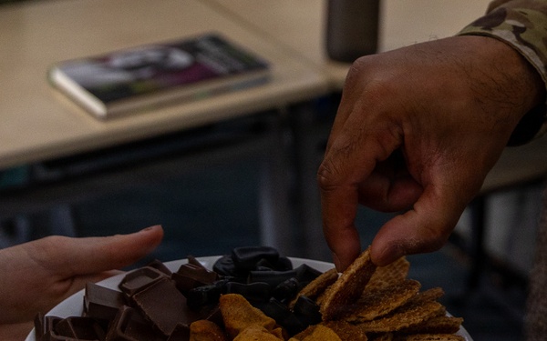 Two people are sharing a plate of assorted snacks, with one person holding the plate and another reaching to take a chip. The plate contains a variety of snacks including chocolate pieces, crackers or waffle chips, and what appear to be cheese puffs or similar orange snacks. One individual is wearing a military camouflage uniform sleeve, visible in the upper right of the frame.