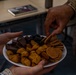 Two people are sharing a plate of assorted snacks, with one person holding the plate and another reaching to take a chip. The plate contains a variety of snacks including chocolate pieces, crackers or waffle chips, and what appear to be cheese puffs or similar orange snacks. One individual is wearing a military camouflage uniform sleeve, visible in the upper right of the frame.